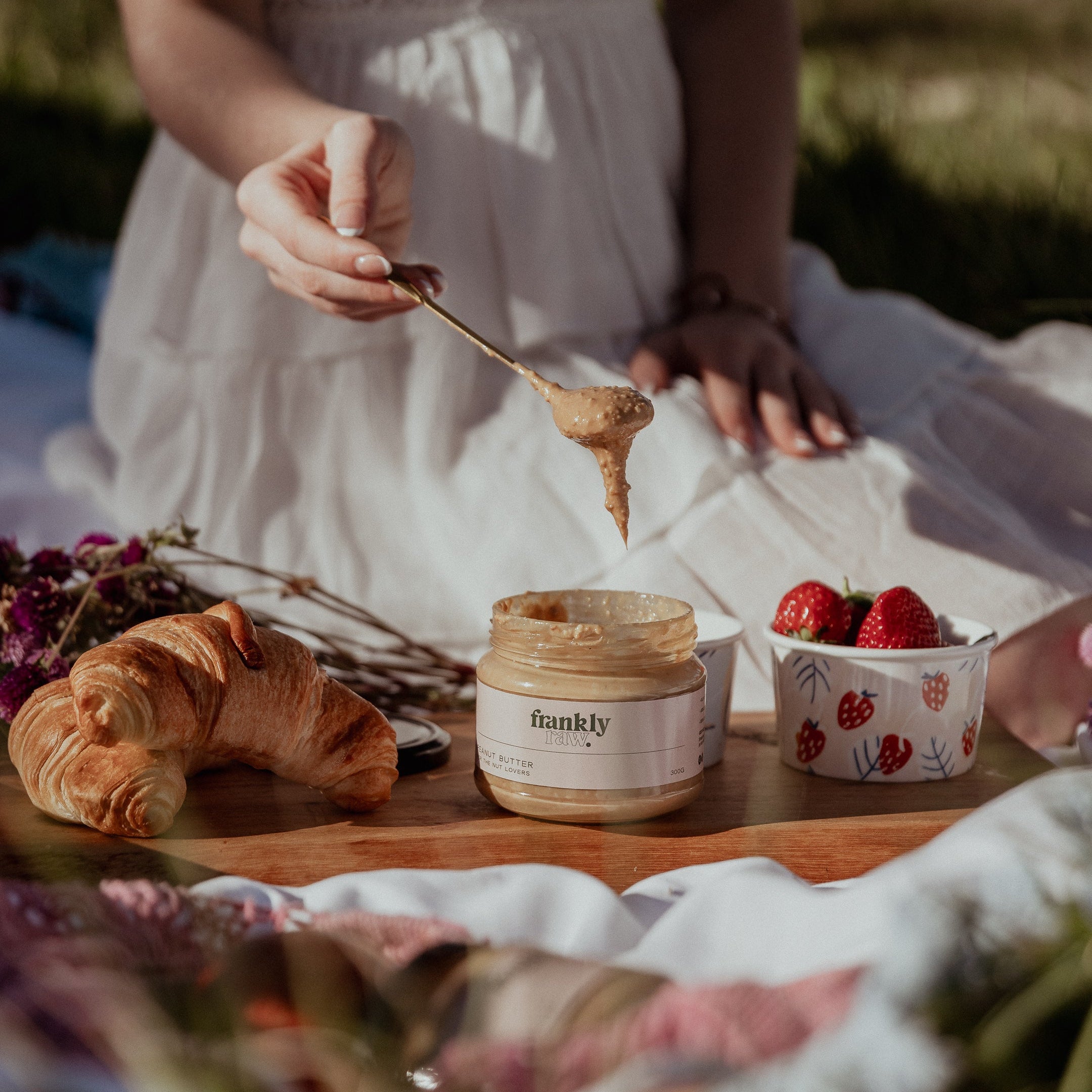 Person in a white dress holding a spoon over a jar of Baskly peanut butter with a croissant and strawberries on a wooden board.