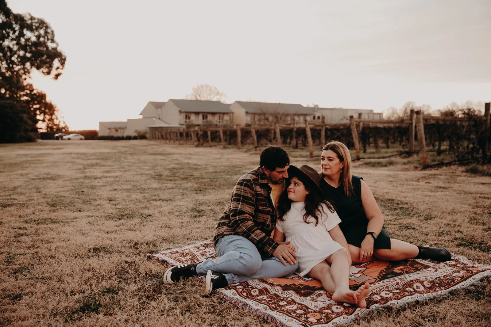 Family of three sitting on a blanket in a field with a building in the background