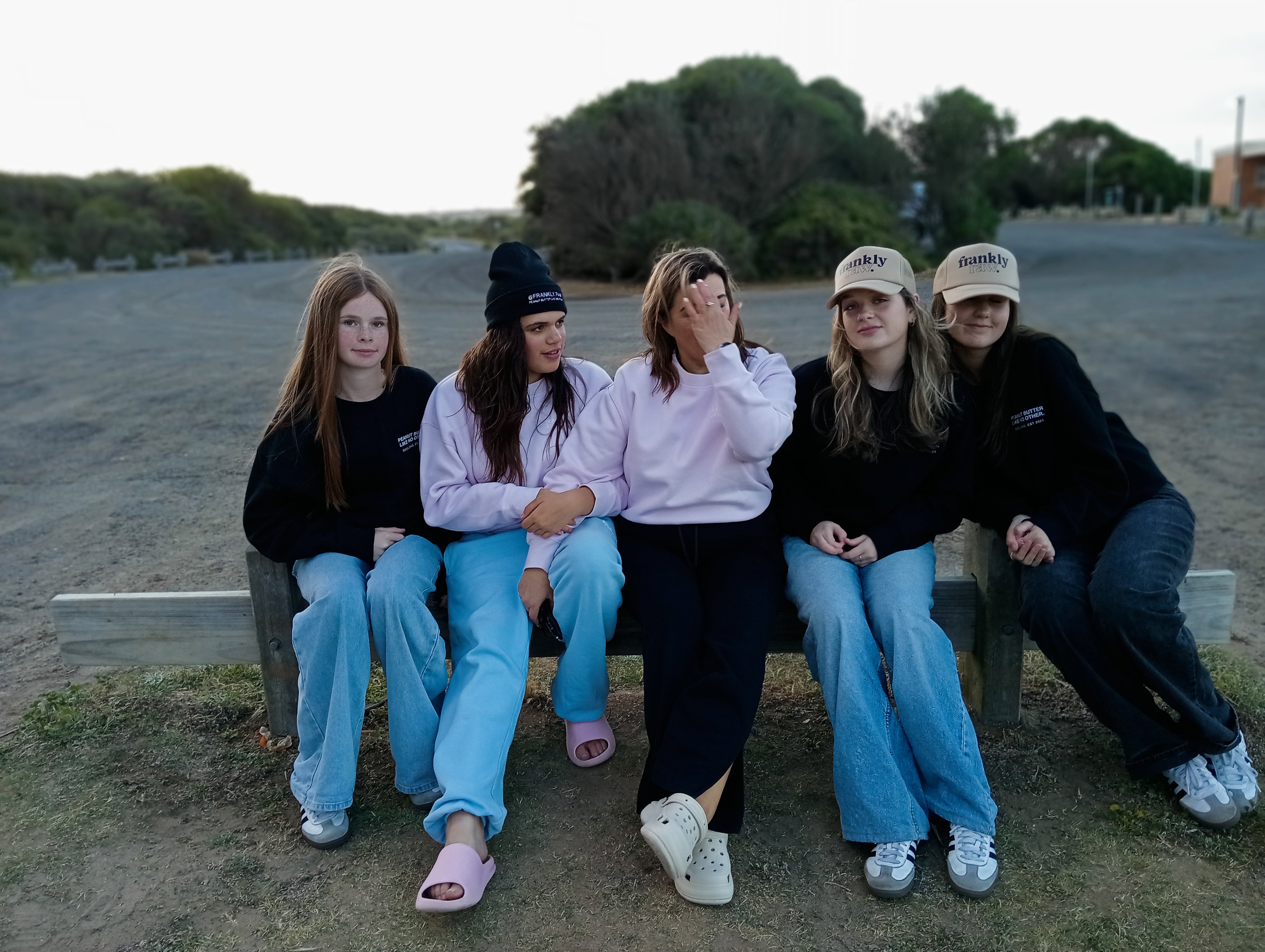 Five young women sitting on a bench outdoors with trees in the background