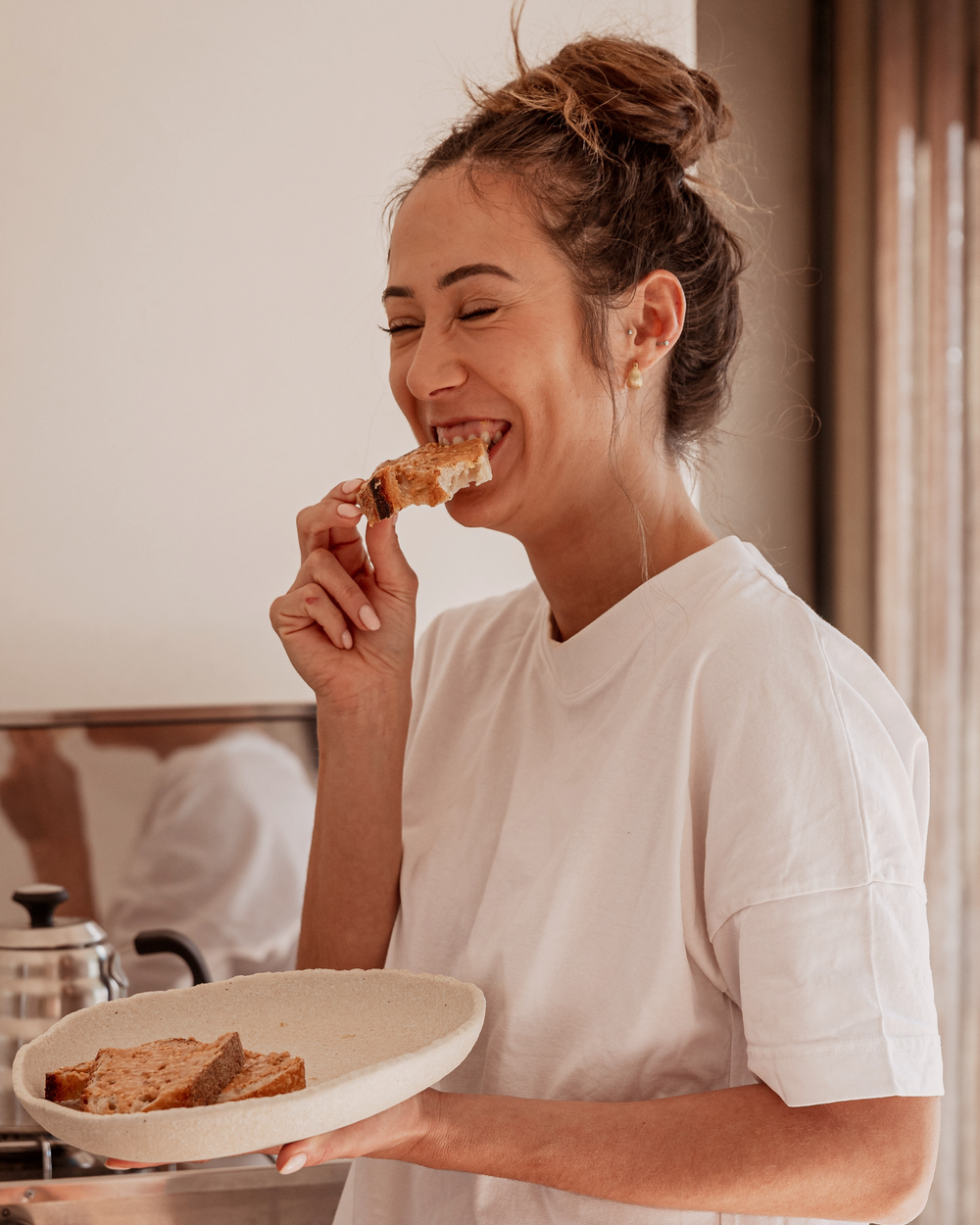 Woman eating a piece of bread while holding a plate with more bread in a kitchen setting.